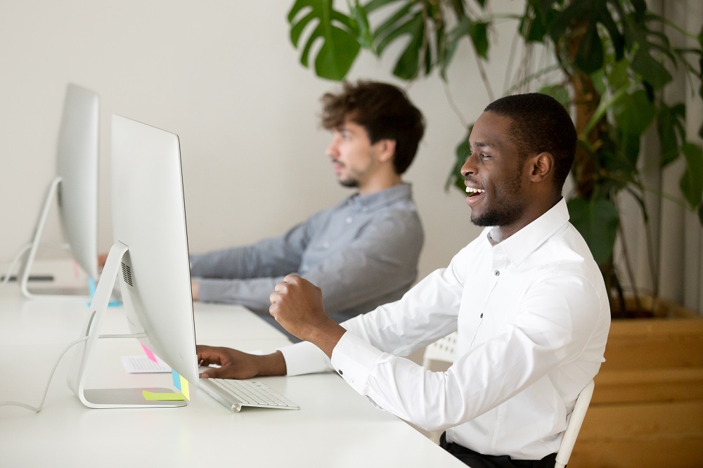 Happy black employee excited by win looking at computer watching game online in office, african winner celebrating good result success achievement, motivated man reading great news being promoted